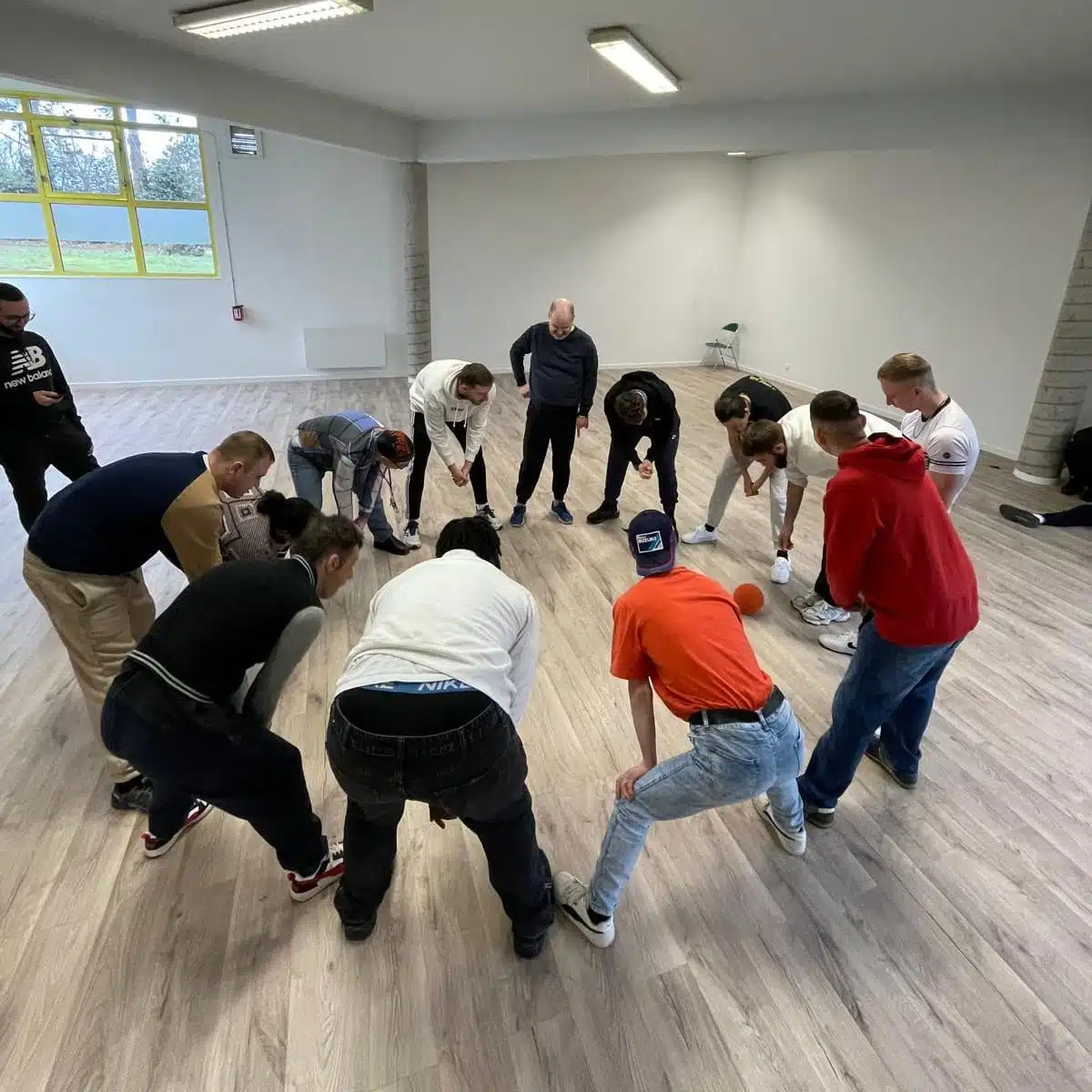Un groupe de 13 personnes en BPJEPS participe à une activité dans une salle lumineuse avec un sol en bois. Une balle se trouve au milieu, les participants sont tous penchés en avant et doivent toucher la balle avec leur mains.