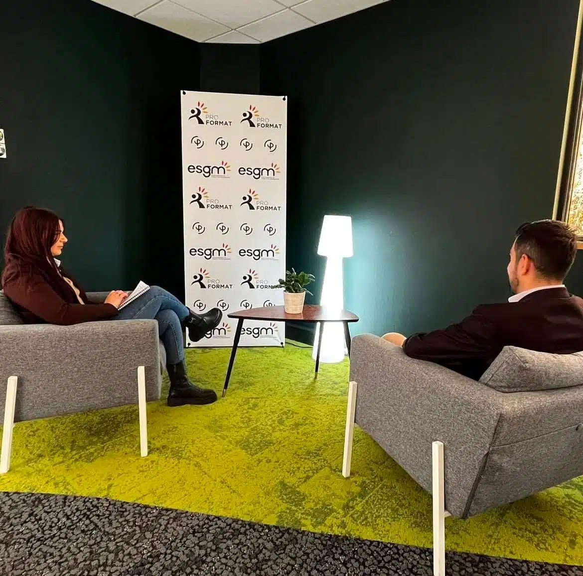 Une femme et un homme discutent dans un espace professionnel. Ils sont assis sur des fauteuils gris, séparés par une petite table en bois avec une plante. En arrière-plan, un kakémono avec les logos de Pro Format et ESGM, et une lampe blanche éclaire la scène.