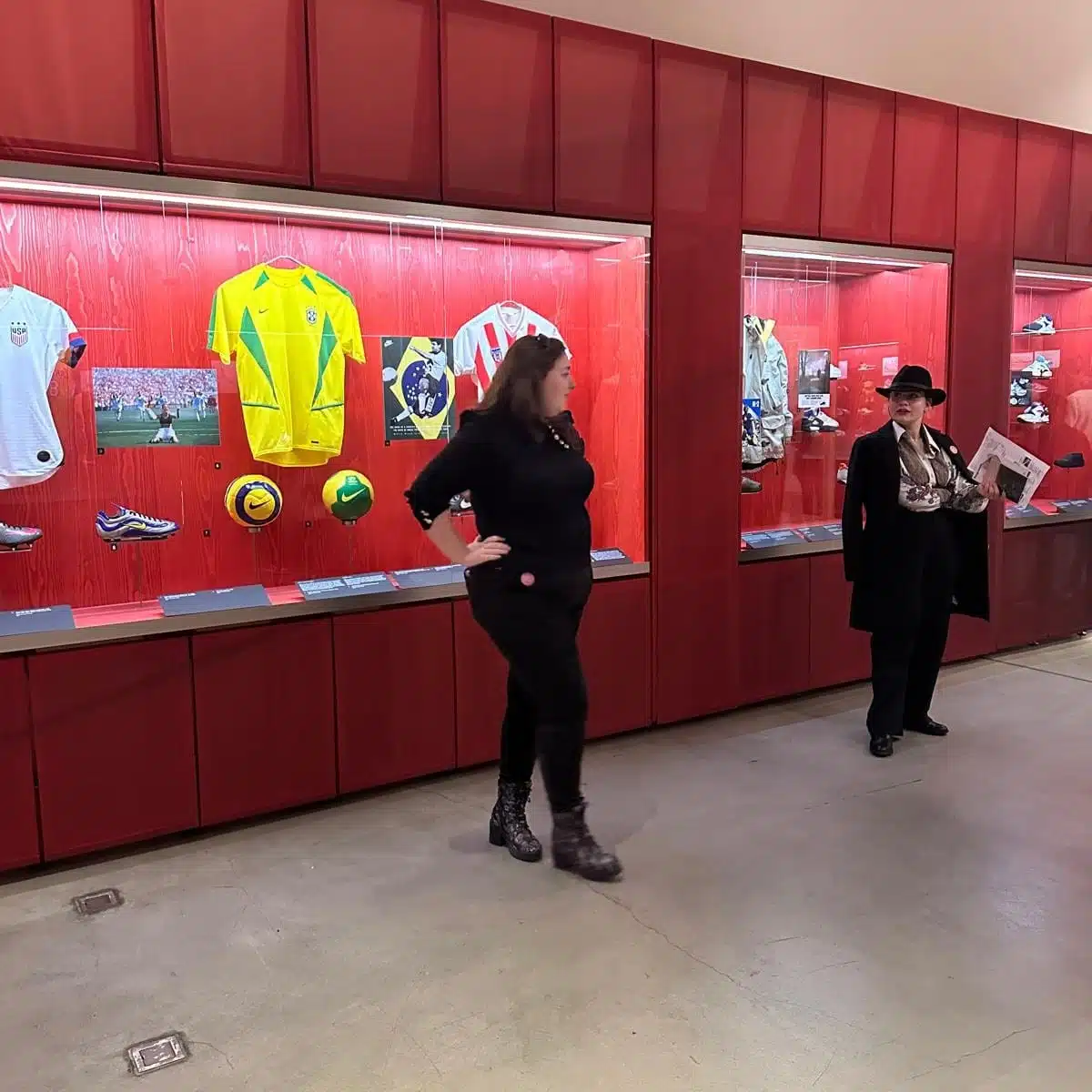 Une guide et une visiteuse explorent un espace d'exposition. En arrière-plan, des vitrines rouges contenant des maillots de football, des ballons et des chaussures, évoquant une thématique sportive.