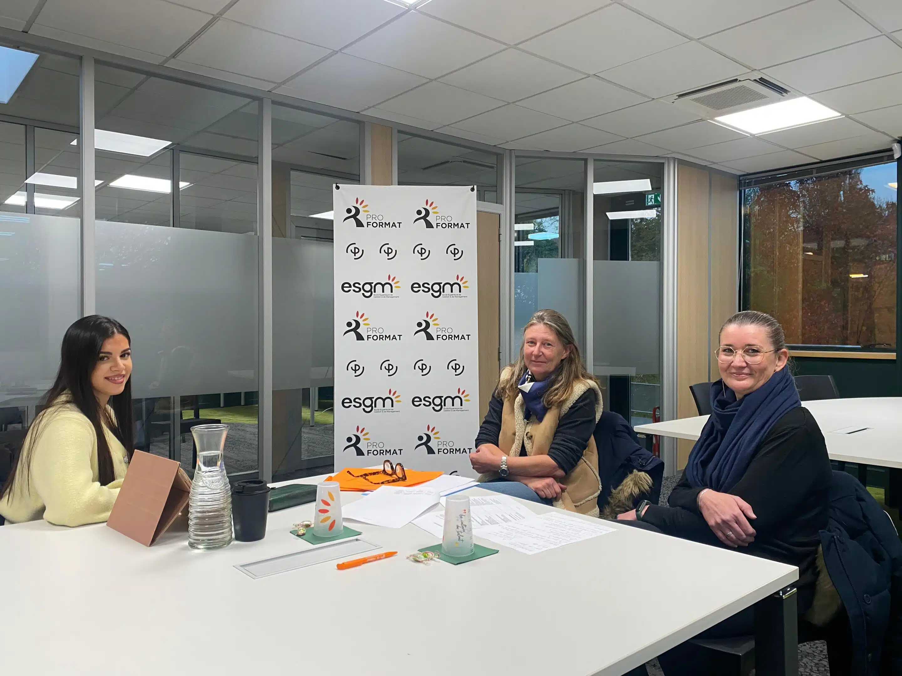 Trois femmes sont assises autour d’une table dans une salle de réunion lumineuse. À gauche, une femme aux cheveux longs et foncés sourit vers l’appareil. Sur la table se trouvent une carafe d’eau, une tasse, des documents et du matériel de travail. Au centre, un panneau affiche les logos d’ESGM et ProFormat. Les deux autres femmes, assises à droite, regardent également vers la caméra. La pièce possède de grandes parois vitrées et un éclairage au plafond.